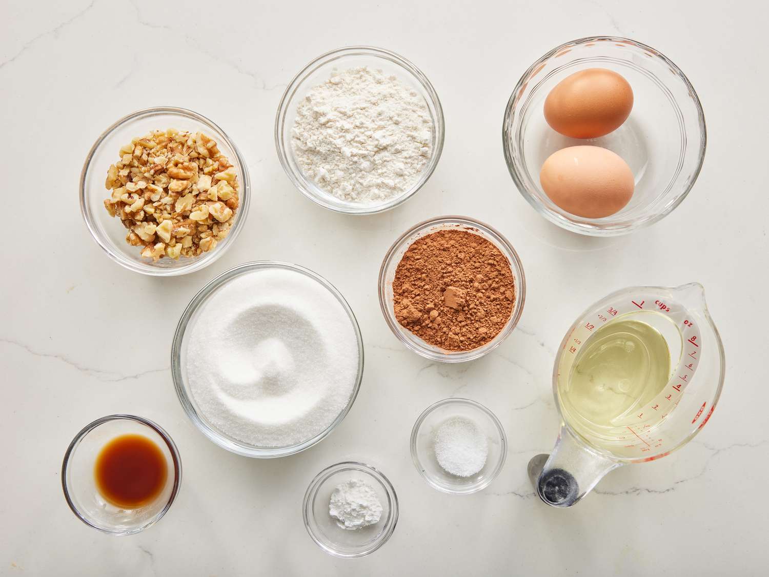 an overhead view of all ingredients to make brownies gathered in small glass bowls on a marble countertop.
