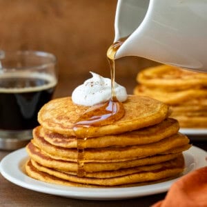 Drizzling syrup over Sweet Potato Pancakes on a white plate on a wooden table.